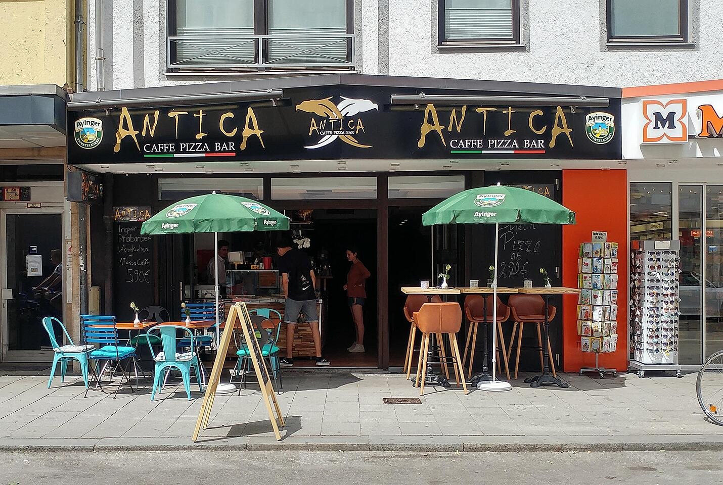 Die Ladenfront von Antica: schwarzes Schild mit goldenem Schriftzug, grüne Sonnenschirme, Tische und Stühle auf dem Gehweg